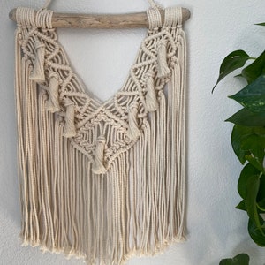 May include: A macrame wall hanging with a natural wood dowel and a geometric pattern. The hanging has a fringe of tassels and is made with cream-colored yarn.