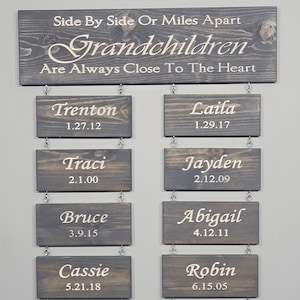 May include: A gray wooden sign with the text "Side By Side Or Miles Apart Grandchildren Are Always Close To The Heart".  The sign has a series of smaller gray wooden signs hanging from it, each with a name and date. The names are Trenton, Laila, Traci, Jayden, Bruce, Abigail, Cassie, Robin, Ethan, Ryan, James, and Lyric.