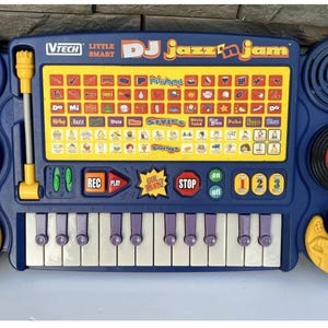 May include: A blue VTech Little Smart DJ Jazz 'n Jam electronic musical toy featuring a central control panel with a grid of colorful buttons, a keyboard with white and purple keys, a green knob, a red knob, a yellow lever, and a black turntable.