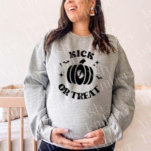 Può includere: Una felpa grigio melange con una grafica nera che recita "Kick or Treat". La grafica presenta una zucca con impronte di piedi di bambino all'interno. Il design include anche pipistrelli e stelle, perfetto per Halloween o un baby shower.