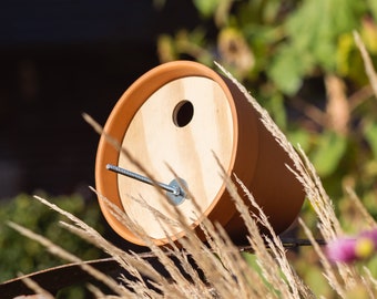 May include: A terracotta pot birdhouse with a wooden front and a small hole for birds to enter. The birdhouse is mounted on a metal stand and is surrounded by tall, brown grass.