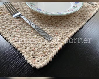 Op de afbeelding: Een gehaakt placemat in crèmekleur met een subtiel gespikkeld patroon. Een zilveren vork en een wit bord met een bloemenpatroon liggen op de placemat.