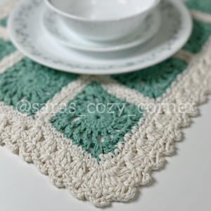 May include: A crocheted placemat with a white border and a green center. The placemat is made with a square pattern and has a scalloped edge.