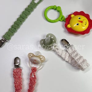 May include: Four crocheted pacifier clips with different colors and designs. One clip is green, one is white, one is pink, and one is attached to a yellow lion toy with a green ring.