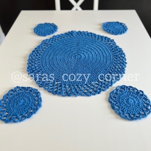Peut inclure: Un ensemble de quatre sets de table en crochet bleu, un grand et trois plus petits, sur une table blanche. Les sets de table ont un design délicat et ajouré.