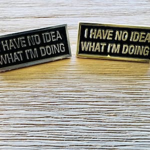 May include: Two pins with the text "I have no idea what I'm doing" on them. One pin is silver and the other is gold.