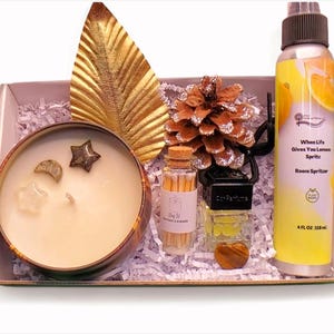 May include: A gift box with a candle, room spritzer, matches, and decorative items. The candle has star and moon-shaped crystals. The room spritzer is yellow with the text "When Life Gives You Lemon Spritz". The box contains a gold leaf and a pinecone.
