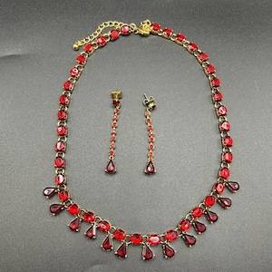 May include: A gold-toned necklace and earring set featuring vibrant red gemstones. The necklace has a series of oval-shaped stones and teardrop pendants. The earrings are long, dangling, and adorned with matching red gems.