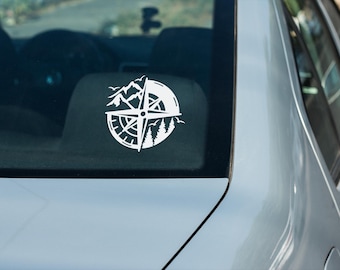 Puede incluir: Calcomanía de coche de vinilo blanco con un diseño de rosa de los vientos con montañas, árboles y pájaros. La calcomanía está aplicada en la ventana trasera de un coche plateado. El diseño es de forma circular.