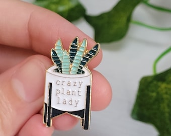 Puede incluir: Un pin de esmalte dorado con una maceta blanca con un diseño de planta verde y negra. El pin dice "crazy plant lady" en texto negro. El pin se sostiene en una mano.