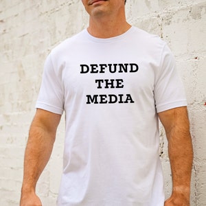 May include: White t-shirt with the text "DEFUND THE MEDIA" printed in black. The shirt is short-sleeved and appears to be made of cotton. The person wearing the shirt is standing in front of a white brick wall.
