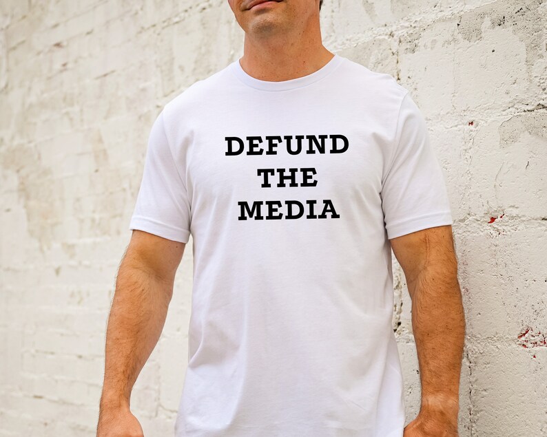 May include: White t-shirt with the text "DEFUND THE MEDIA" printed in black. The shirt is short-sleeved and appears to be made of cotton. The person wearing the shirt is standing in front of a white brick wall.