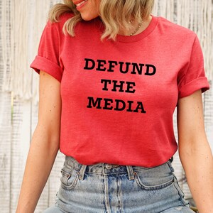 May include: A red t-shirt with the text "DEFUND THE MEDIA" in black block letters. The shirt has rolled-up sleeves and is paired with blue denim jeans. The overall style is casual and comfortable.