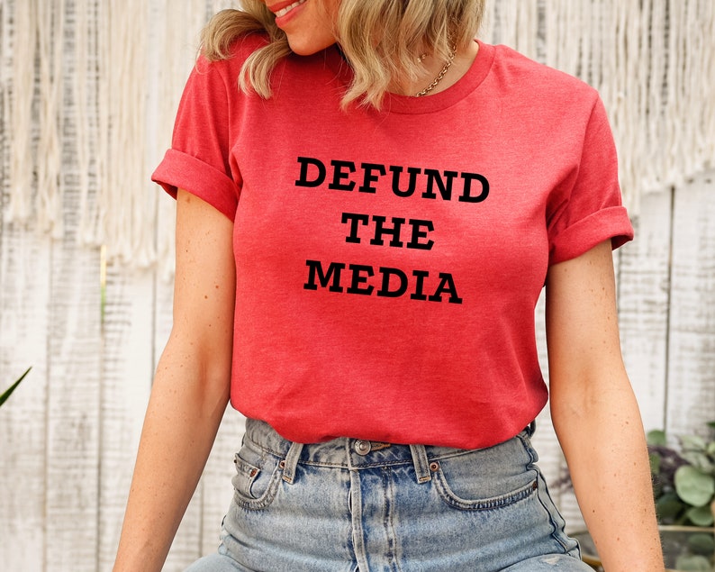 May include: A red t-shirt with the text "DEFUND THE MEDIA" in black block letters. The shirt has rolled-up sleeves and is paired with blue denim jeans. The overall style is casual and comfortable.