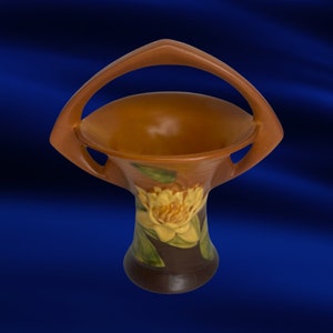 May include: A brown ceramic basket with a handle. The basket features a floral design with yellow and green accents. The handle is arched, and the basket has a wide opening. The base is dark brown.