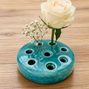 May include: A round, teal ceramic flower frog with multiple circular openings for stems. A single cream-colored rose and a small sprig of white baby's breath are inserted into two of the holes, showcasing the functional design of this glazed pottery piece.
