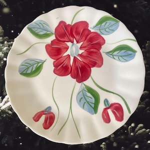 May include: A white ceramic plate with a scalloped edge, hand-painted with a large red flower and green and blue leaves. The floral design is centered on the plate, with additional red buds and leaves around the edges. A decorative piece.