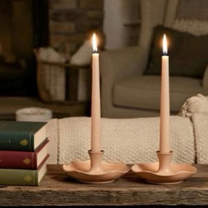 May include: Two lit, pale pink taper candles in matching holders. The holders have a scalloped edge and are sitting on a wooden surface. A stack of books is visible in the background, suggesting a cozy, home decor theme.