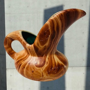 May include: A brown and tan ceramic pitcher with a unique, curved spout and a handle. The interior is a deep green. The pitcher has a marbled appearance, with swirling patterns of light and dark brown. The pitcher is likely used for serving beverages.