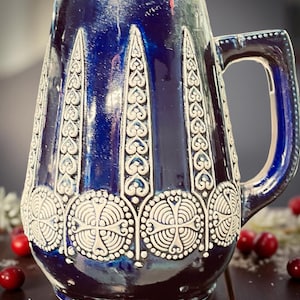 May include: A dark blue ceramic beer stein with a handle. The stein features white, ornate, raised relief designs. The designs include vertical patterns and circular patterns with crosses. The stein is on a dark wooden surface.