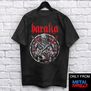 Puede incluir: Camiseta negra con un gráfico de una calavera con cuchillas cruzadas y la palabra "haraka" en letras góticas rojas. El diseño está dentro de un emblema circular. La camiseta está colgada en una percha negra.