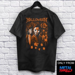 Könnte beinhalten: Schwarzes T-Shirt mit einer orangefarbenen Grafik von Michael Myers aus der Halloween-Filmreihe. Die Grafik zeigt eine gruselige Waldszene mit Vollmond und fallenden Blättern. Der Text "HALLOWEEN" ist über der Grafik gedruckt.