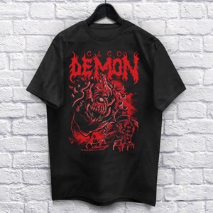 以下が含まれることがあります： 赤い悪魔のグラフィックが描かれた黒いTシャツ。上には「CACO DEMON」の文字が書かれている。