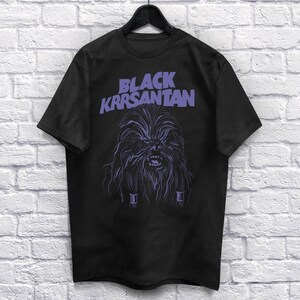 Op de afbeelding: Zwart T-shirt met een paarse illustratie van een Wookiee-achtig personage en de tekst "BLACK KRRSANTAN". Het ontwerp heeft gedetailleerde vacht- en gezichtskenmerken, wat een opvallende uitstraling cre&euml;ert.