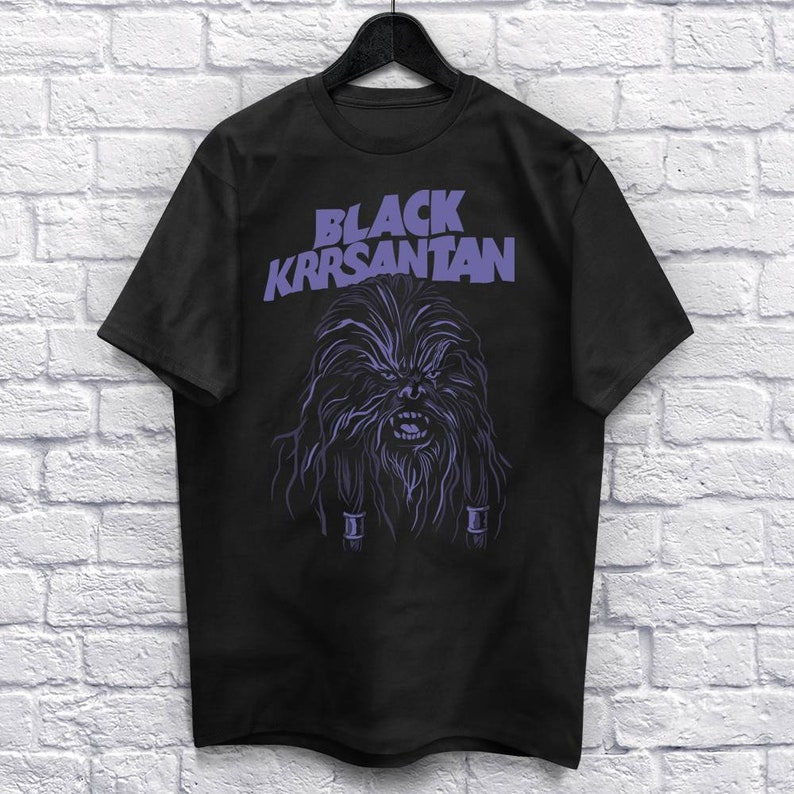 Op de afbeelding: Zwart T-shirt met een paarse illustratie van een Wookiee-achtig personage en de tekst "BLACK KRRSANTAN". Het ontwerp heeft gedetailleerde vacht- en gezichtskenmerken, wat een opvallende uitstraling cre&euml;ert.