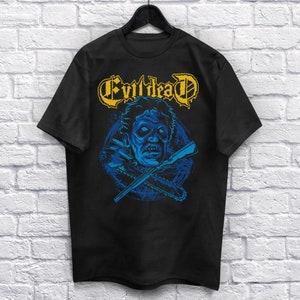 May include: Black t-shirt featuring a blue and yellow graphic of a zombie head with a chainsaw and a rifle. The text "Evil Dead" is written in yellow above the graphic.