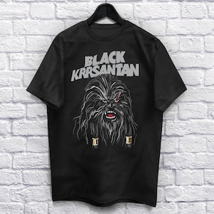 Op de afbeelding: Zwart T-shirt met een afbeelding van een Wookiee-achtig personage en de tekst "BLACK KRRSANTAN" in grijs. Het personage heeft lang, donker haar, een litteken en gouden accenten. Het T-shirt hangt aan een zwarte hanger.