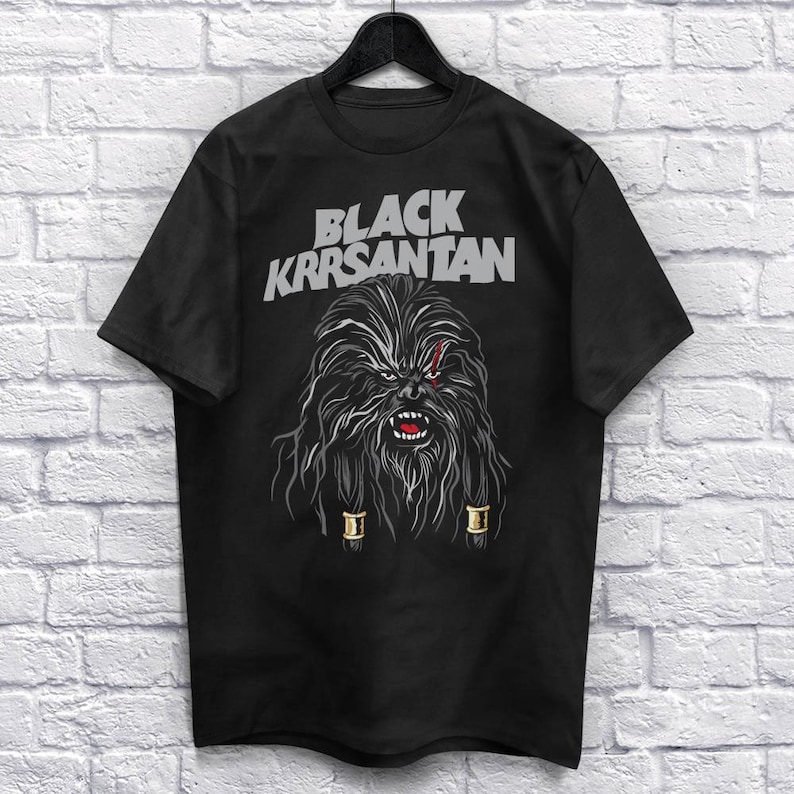Op de afbeelding: Zwart T-shirt met een afbeelding van een Wookiee-achtig personage en de tekst "BLACK KRRSANTAN" in grijs. Het personage heeft lang, donker haar, een litteken en gouden accenten. Het T-shirt hangt aan een zwarte hanger.