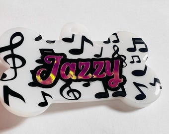 May include: A white, bone-shaped pet tag with black musical notes and the name "Jazzy" in a pink and gold iridescent font. The tag is designed for a pet and features a musical theme.