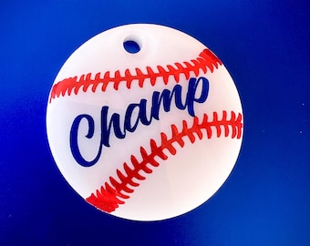 Puede incluir: Adorno de béisbol blanco con costuras rojas y la palabra "Champ" en letras azules.