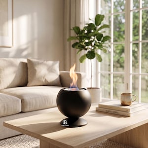 May include: A black, spherical tabletop fireplace with a flame. The fireplace sits on a wooden table in a living room setting. The flame is orange and yellow. A beige sofa, plant, and books are in the background.