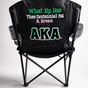 May include: Black folding chair with a black frame and mesh cup holder. The back of the chair features green and pink text that reads "What Up Doe Thee Centennial 146 A. Green AKA".