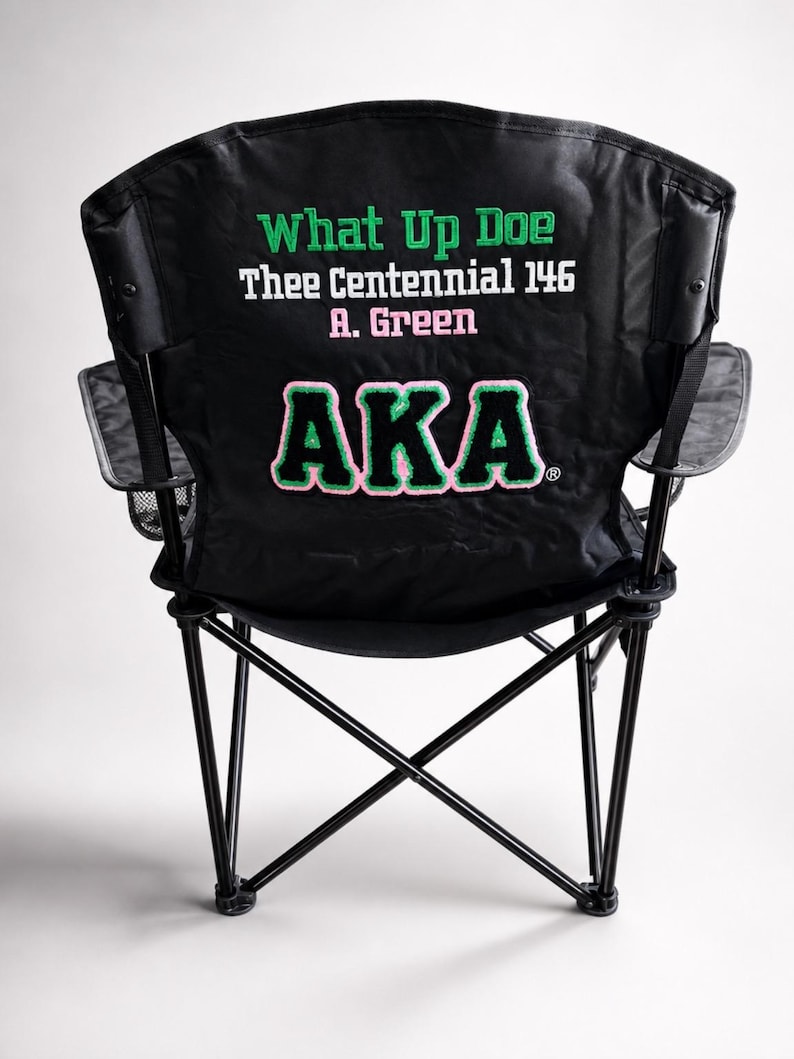 May include: Black folding chair with a black frame and mesh cup holder. The back of the chair features green and pink text that reads "What Up Doe Thee Centennial 146 A. Green AKA".