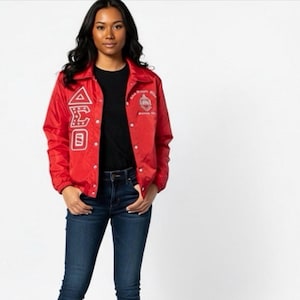 May include: A red nylon jacket with white embroidered Greek letters and a crest on the front. The jacket has a button-up closure and a collar. The person is wearing a black shirt and blue jeans.
