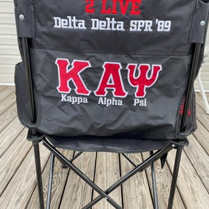 May include: Black folding chair with red and white embroidered text. The text includes "2 LIVE", "Delta Delta SPR '89", and "KAY" with "Kappa Alpha Psi" below. The chair has a black metal frame and a black fabric seat and back.