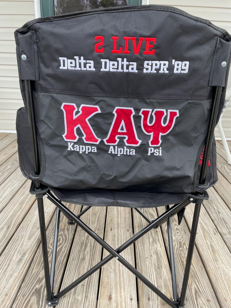 May include: Black folding chair with red and white embroidered text. The text includes "2 LIVE", "Delta Delta SPR '89", and "KAY" with "Kappa Alpha Psi" below. The chair has a black metal frame and a black fabric seat and back.