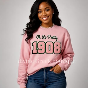 May include: A light pink long-sleeve crewneck sweatshirt featuring the text Oh So Pretty in dark green script above the large, textured, light pink numbers 1908, which are outlined in dark green. The sweatshirt is paired with blue denim jeans.