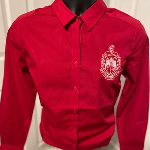 May include: A red long-sleeved button-down shirt with a white embroidered crest featuring the Greek letters "Alpha Sigma Beta".