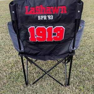 May include: Black folding camping chair with the name "LaShawn" embroidered in red, along with "SPR '93" and the number "1913". The chair has a black frame and is set on a grassy surface.