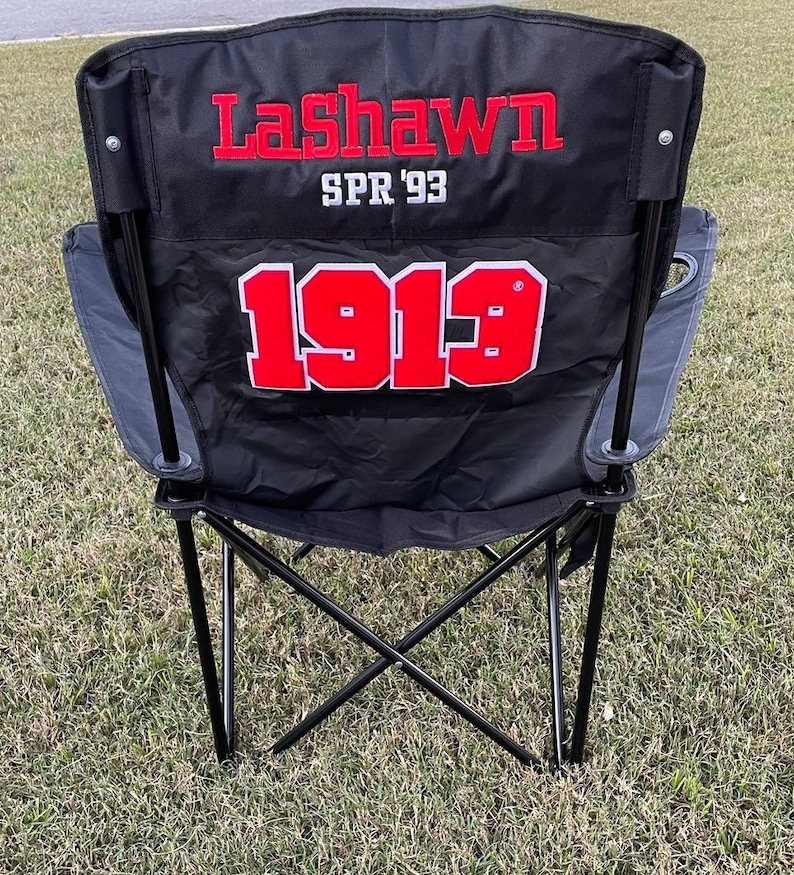 May include: Black folding camping chair with the name "LaShawn" embroidered in red, along with "SPR '93" and the number "1913". The chair has a black frame and is set on a grassy surface.