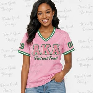 May include: A pink V-neck t-shirt with green and white striped trim on the sleeves and neckline. The front features the green and pink text AKA and First and Finest, with the number 08 on the sleeves. The shirt is paired with blue denim jeans.