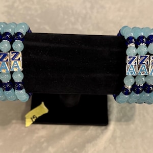 May include: Two beaded bracelets with light blue and dark blue beads. Each bracelet has silver lettered charms spelling out "ZZZZZ" and "AAAAA". The bracelets are displayed on a black velvet stand.