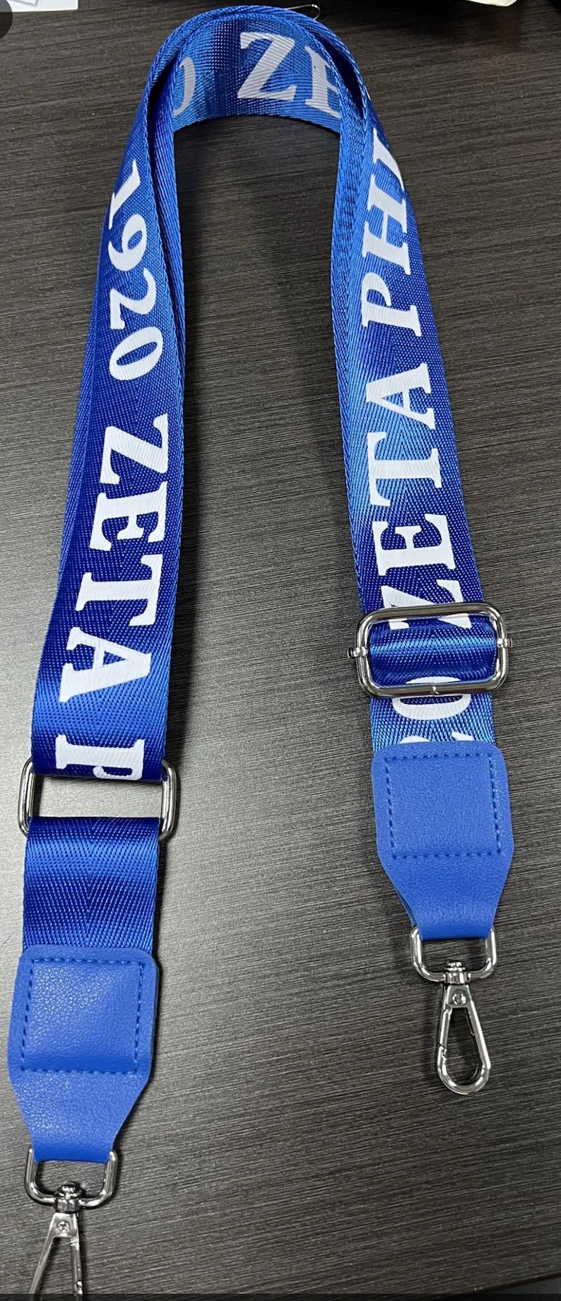 Puede incluir: Lanyard azul y blanco con el texto "Zeta Phi 1920" impreso en &eacute;l. El lanyard tiene un clip plateado en cada extremo.
