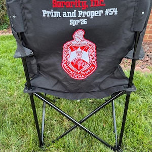 May include: Black folding chair with the text "Delta Sigma Theta Sorority, Inc. Prim and Proper #54 Spr'26" in red and white. The chair has a red and white emblem on the back.