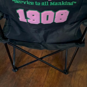May include: Black folding chair with the text "Alpha Kappa Alpha Sorority, Inc." in pink and green. The text "Service to all Mankind" is also visible. The number "1908" is displayed in pink with a green border.