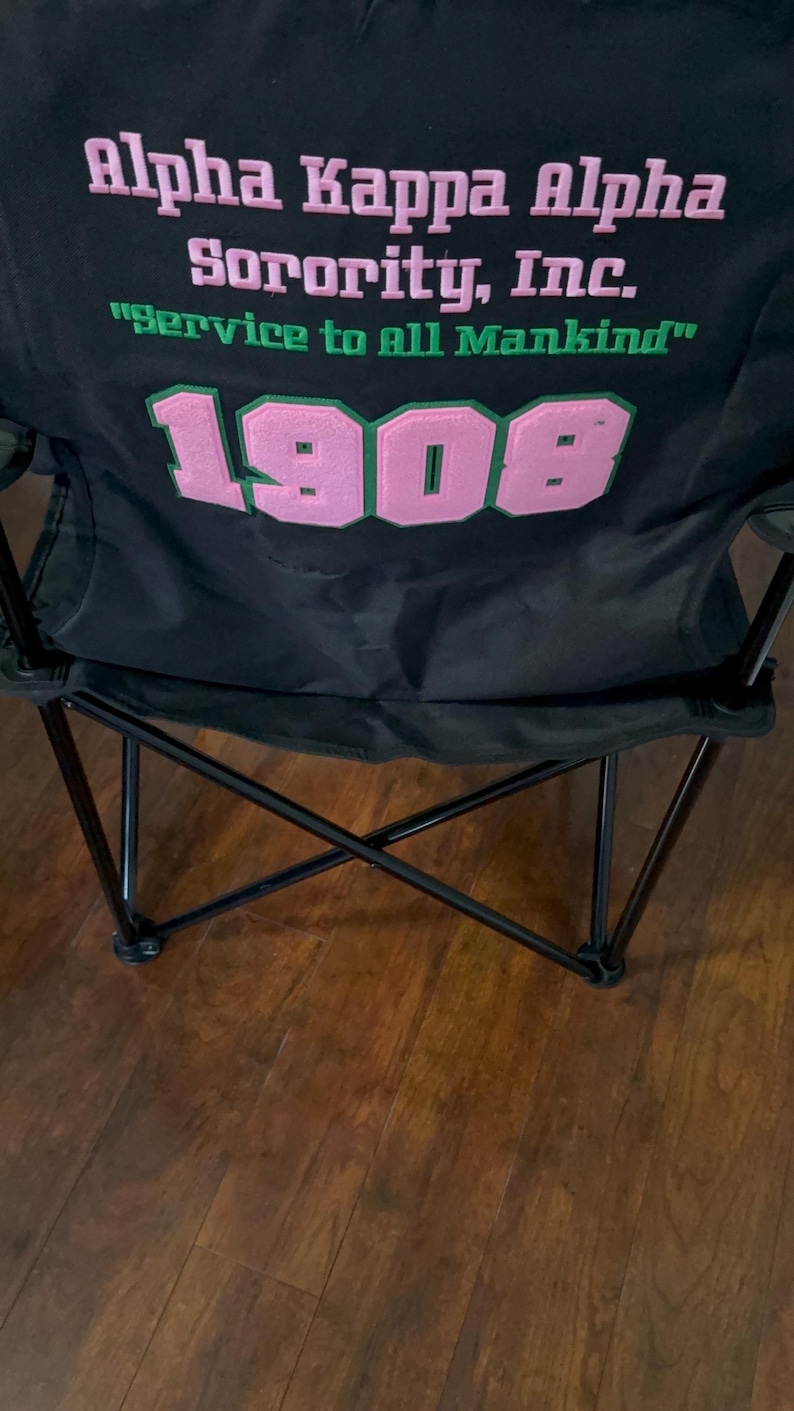 May include: Black folding chair with the text "Alpha Kappa Alpha Sorority, Inc." in pink and green. The text "Service to all Mankind" is also visible. The number "1908" is displayed in pink with a green border.
